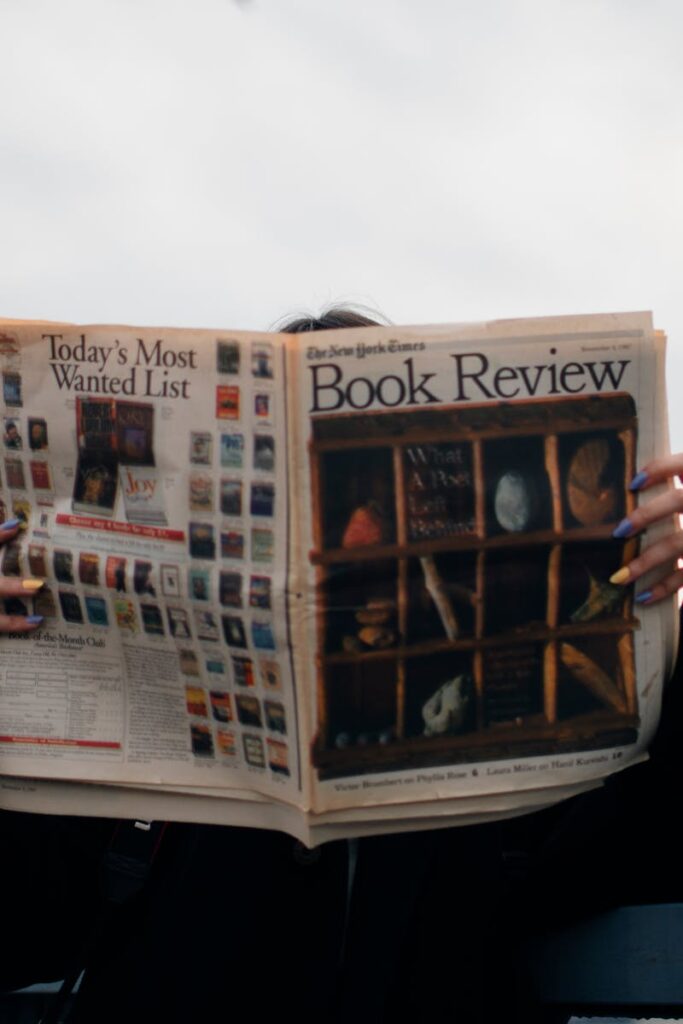 Individual holding a book review newspaper with colorful nails against a cloudy sky backdrop.