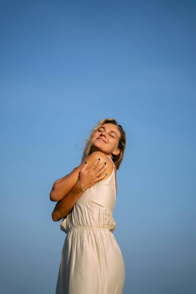 Woman in white dress embracing sunlight with closed eyes against blue sky.