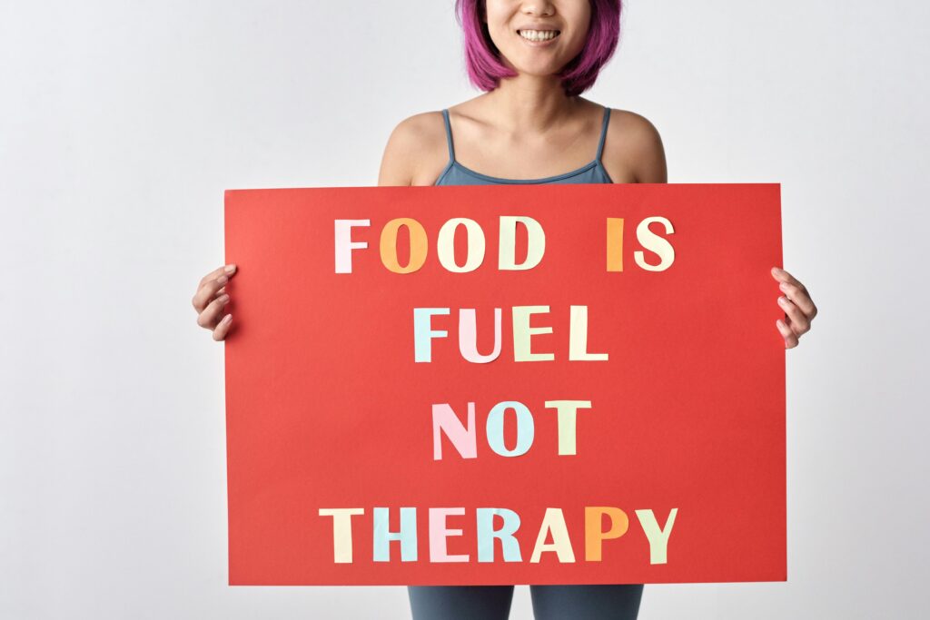 A woman with pink hair holding a red sign reading 'Food is Fuel Not Therapy' on a white background.